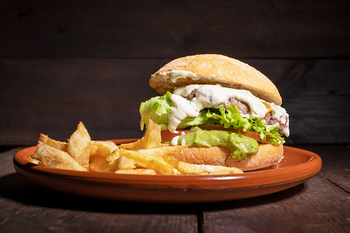 Delicious gourmet beef burger with creamy sauce, lettuce, tomato and guacamole, served with french fries on a rustic plate on a wooden table. Tracking shot. High quality photography.