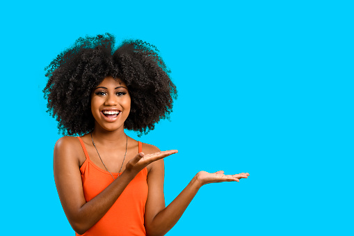 a young black woman with afro hairstyle smiles and points with her hands to the right side, a space for your product or message, person, advertising concept, isolated or blue background.