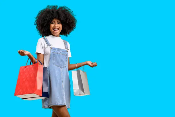 Photo of Portrait of a smiling young girl with dark skin holding shopping bags, isolated on a blue background.