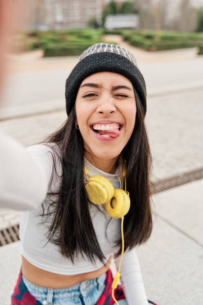 라티나 그녀의 혀를 고집 재미있는 얼굴을만드는 동안 셀카를 복용. 캐주얼 한 옷을 입고 헤드폰미소모자를 쓰고 있는 젊은 히스패닉 소녀. - humor music headphones women 뉴스 사진 이미지