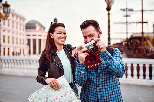 Smiling Male Tourist Showing Girlfriend Different Photos Of Landmarks While Visiting City As Tourists