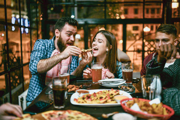 lächelndes paar füttert sich gegenseitig mit pizza, während es mit freunden im restaurant zu abend isst - city chicken stock-fotos und bilder