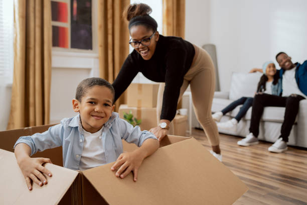 mom playing with baby, pushing a cardboard box across the living room floor where her sweet little son sits, joy at moving into a new apartment, smiling boy - family large american culture fun imagens e fotografias de stock