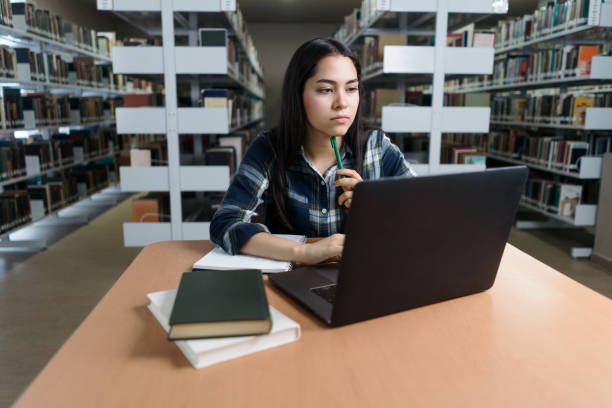 istock Female student holding pencil and using laptop in library 1372604886