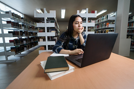 A latin female student using a laptop computer at a table in the library.