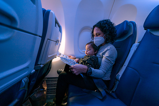 A young mom of Pacific Islander descent wearing a protective face mask holds her Eurasian toddler daughter in her lap and reads her a book while riding on an airplane.