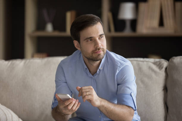 istock Pensive positive young man using mobile phone, sitting on couch 1372613207
