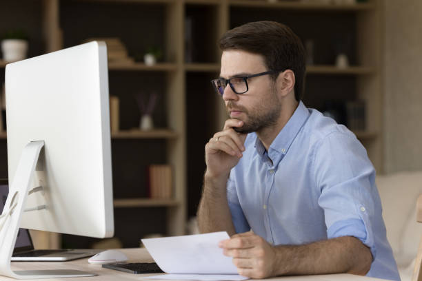 Serious thoughtful young business man in glasses working at monitor Serious thoughtful young business man in glasses working at monitor in office, using computer at workplace, thinking over paper report, touching chin, watching, reading content on display giant cube stock pictures, royalty-free photos & images