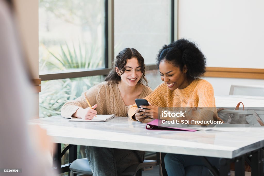 Teenage girls look at smartphone during class Two female high school friends look at something on a smartphone before class begins. Laughing Stock Photo
