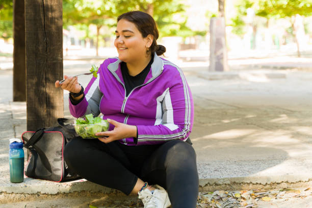 Beautiful active woman enjoying a salad Happy overweight woman smiling while eating a healthy green salad before starting her cardio workout outdoors fat nutrient photos stock pictures, royalty-free photos & images