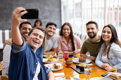 Happy group of Latin American friends eating together at a restaurant and taking a selfie with a cell phone