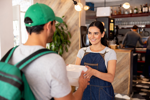 Happy Latin American waitress handling a food order to a delivery person at a dark kitchen