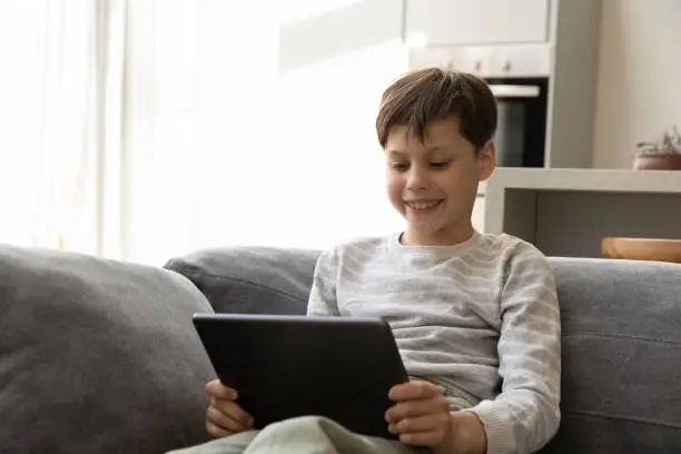 Cheerful smiling gen Z teenage kid watching virtual lesson, online movie, series on tablet computer, playing virtual video game, using digital device, browsing internet, relaxing on couch at home