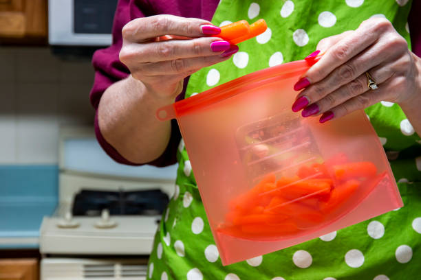A caucasian woman with a green and white polkadot apron is grapping a fresh snack of carrots out of a reusable silicone food baggie. It is a red quart size bag.  The scene takes place in a kitchen.. Zero waste susatainable.  Copy space on left.
