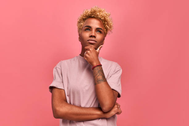 let me think. Thoughtful dark skinned woman with blond haircut, purses lips, keeps hand under chin, plans something or makes choice, makes decision in mind, stands over pink background. Doubt concept