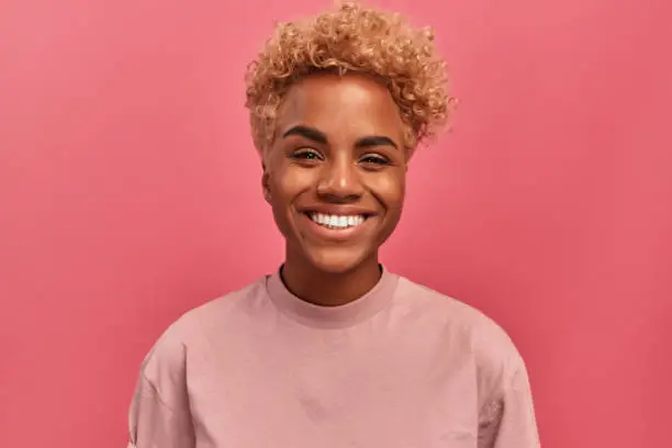 Studio portrait of young dark skinned woman with blonde hair in good mood standing on pink background. Joyful woman posing in studio expressing happiness. Concept of happiness and positive emotions.