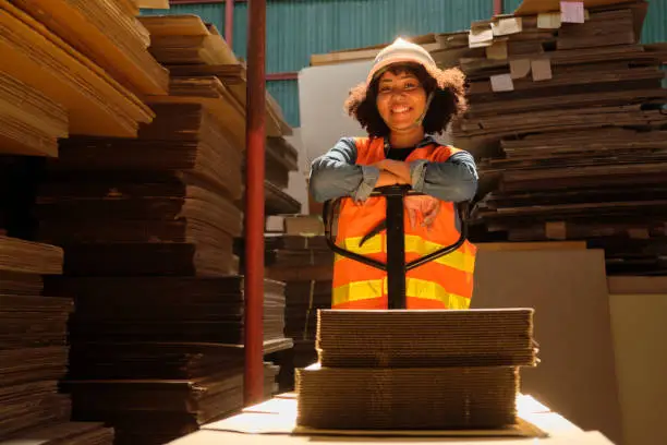 Female warehouse worker in safety uniform and hardhat using hydraulic trolley haul piles of cardboard for shipping and logistic transport at manufacture factory, supply stocks in packaging industrial.