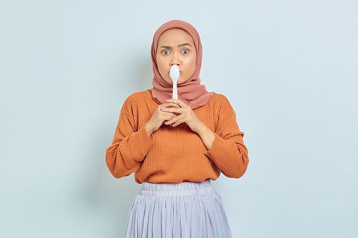 Beautiful Asian woman in brown sweater and hijab covering her mouth with spoon, want to try tasty food with shocked expression isolated over white background