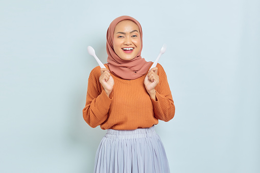 Cheerful beautiful Asian woman in brown sweater and hijab hold fork and spoon before breakfast isolated over white background
