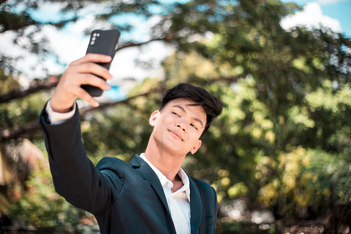 A vain and confident young asian man in business attire takes a selfie of himself while at the park.