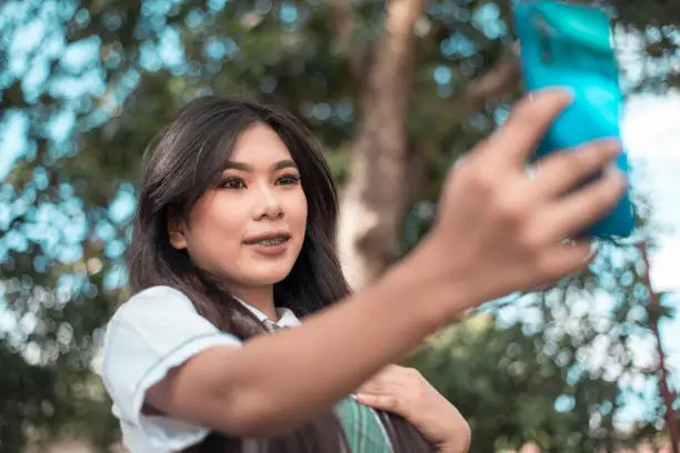 Photo of A pretty vain young asian woman in a student uniform takes a selfie of herself while at the park. Taking a photo for social media.