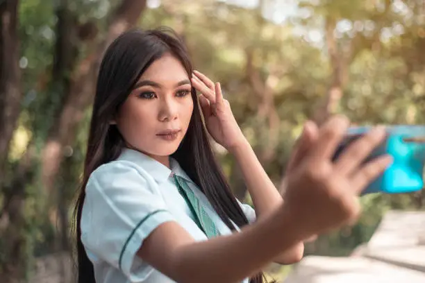 Photo of A pretty vain young asian woman in a student uniform takes a selfie of herself while outdoors. Taking a photo for social media.