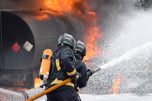 Barcelona, Cataluña, Spain - January 24, 2022: Two firefighters facing a fire and putting it out with a hose, with smoke and heat. They are  equipped with heavy respiratory protection and wears protective equipment that protects them from the heat and flames.