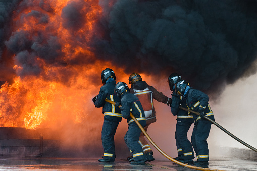 Barcelona, Cataluña, Spain - January 24, 2022: A group of firefighters facing a fire and putting it out with a hose, with smoke and heat. They are  equipped with heavy respiratory protection and wears protective equipment that protects them from the heat and flames.