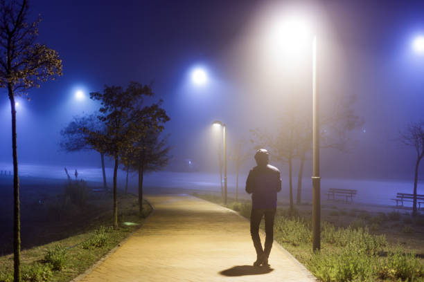 uomo da dietro con un cappello che cammina di notte nella nebbia lungo un marciapiede solitario illuminato da lampioni - bizarre rear view hat blue foto e immagini stock