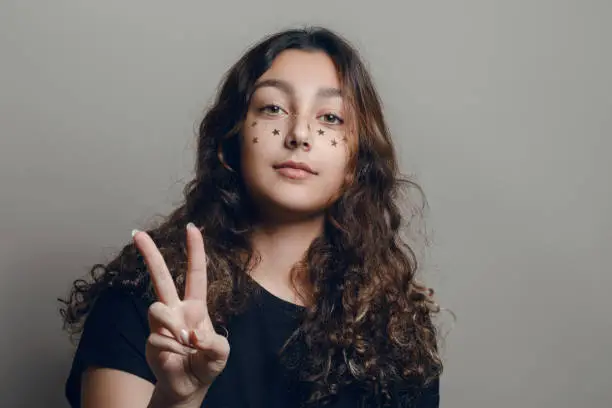 A teenager girl using the gesture as a victory sign. Pretty girl with dark curly hair and sparkles asterisks on her face.
