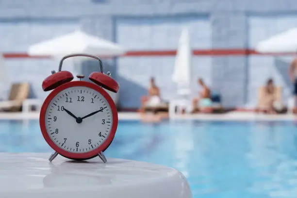 Photo of Red alarm clock standing on edge of hotel swimming pool closeup