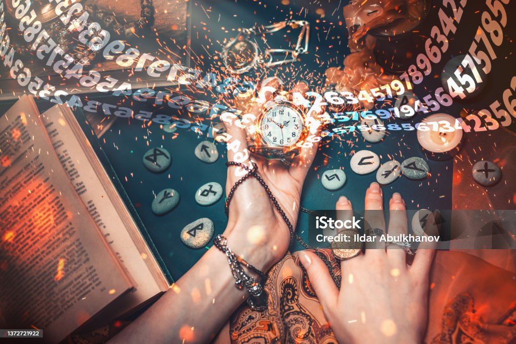 Astrology and esotericism. The female hand of the witch holds a watch on a chain. In the background, old books, fortune-telling runes, candles, and jewels. Sparks fly from the clock and numbers move. Astrology and esotericism. The female hand of the witch holds a watch on a chain. In the background, old books, fortune-telling runes, candles, and jewels. Sparks fly from the clock and numbers move. The view from the top. Numerology Stock Photo