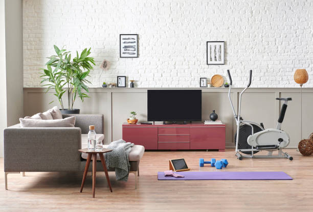 Man is doing sport and looking cell phone training program. Decorative living room, television unit, grey sofa purple mat and blue dumbbell brick wall home interior concept.