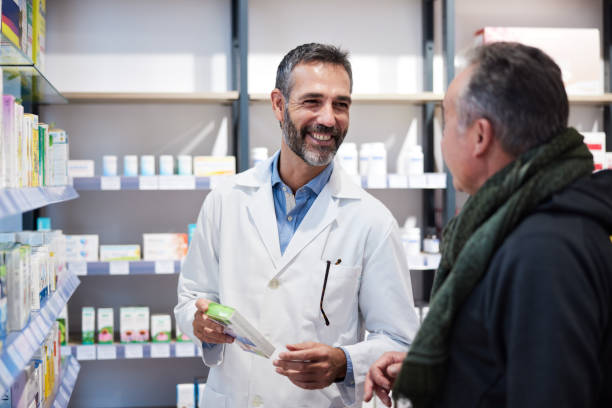 A pharmacist is attending a senior man. A man in a lab coat and a senior man are standing next to a pharmacy shelf. pharmacist stock pictures, royalty-free photos & images