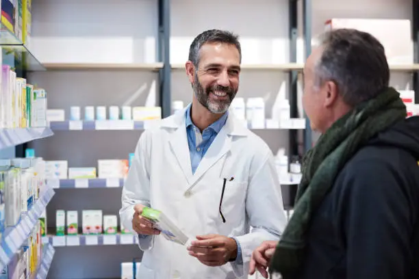 A man in a lab coat and a senior man are standing next to a pharmacy shelf.