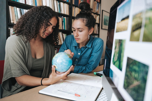 two young woman researching together global issues at desk with computer monitor and globe in hands