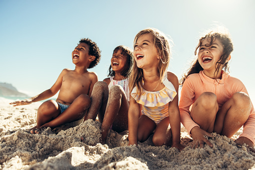 Summer fun at the beach. Four young friends laughing cheerfully while sitting on sea sand at the beach. Group of adorable little kids having a good time together during summer vacation.