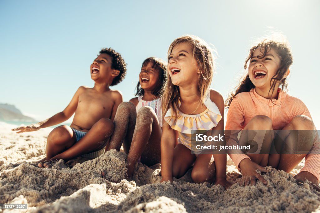 Summer fun at the beach Summer fun at the beach. Four young friends laughing cheerfully while sitting on sea sand at the beach. Group of adorable little kids having a good time together during summer vacation. Child Stock Photo