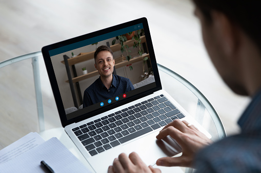 View over businessman freelancer shoulder chatting online by video call, smiling colleague or mentor on laptop screen speaking, young man engaged in internet meeting or interview by webcam