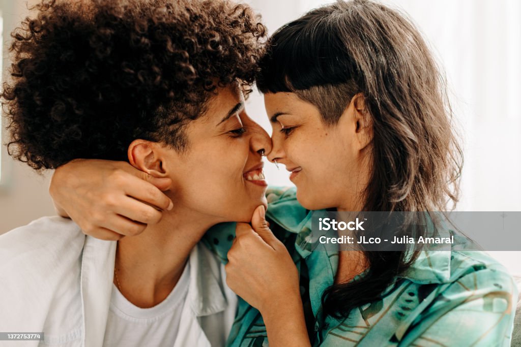 Playful lesbian couple touching their noses together Playful lesbian couple touching their noses together at home. Two affectionate female lovers flirting with each other. Romantic young lesbian couple sitting together in their living room. Couple - Relationship Stock Photo