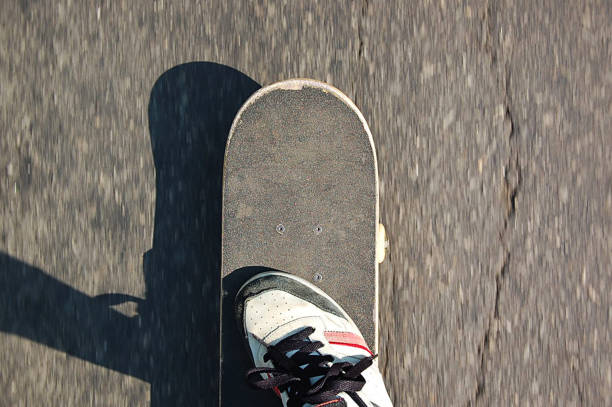 vista superior de um skate e um pé se movendo no asfalto em um dia ensolarado. - skateboarding skateboard teenager extreme sports - fotografias e filmes do acervo