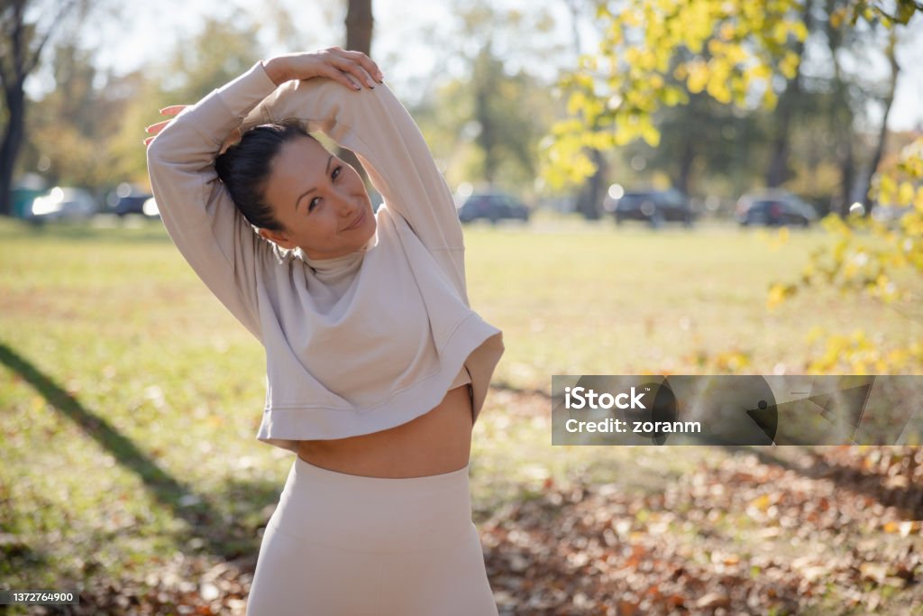 Woman in casual sports clothes stretching arms and smiling at camera, leisure activity outdoors Woman in casual sports clothes stretching arms in public park, holding elbow and bending arm backwards, smiling at camera 35-39 Years Stock Photo