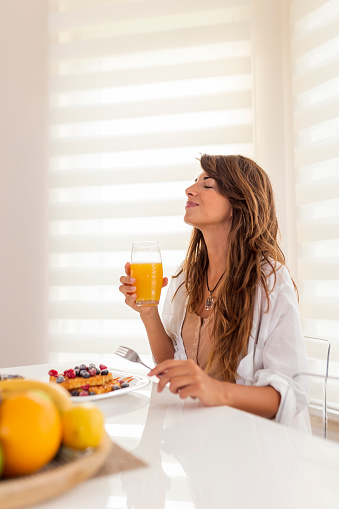 Young woman sitting at dinning room table and smiling, drinking fresh orange juice for breakfast