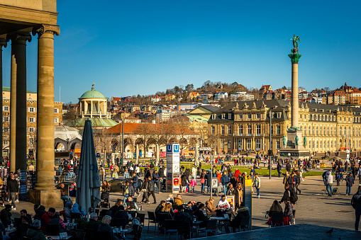 Stuttgart, Germany - February 13, 2022: A crowd of people at town square called 'Schlossplatz' on a sunny day. In background is the obelisk jubilee column and the castle of Stuttgart.
