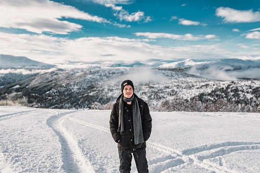 Young adult man standing, looking at camera smiling, in stryn ski resort, norway during winter with plenty of snow, mountain landscape with forets in the distance