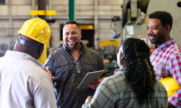 Multiracial group working in factory having meeting A group of four multiracial mature adults, one woman and three men, working in a metal fabrication shop having a meeting on the factory floor. A mixed race Hispanic and Caucasian man is in charge, holding a digital tablet, talking and looking toward an African-American man wearing a hard hat. They are smiling. working class stock pictures, royalty-free photos & images