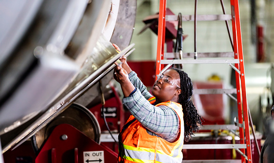 A mature African-American woman working in a metal fabrication shop. She is wearing a safety vest and protective goggles, using a tape measure to measure part of a large metal object.