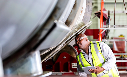 A mature African-American man working in a manufacturing facility specializing in metal fabrication. He is wearing safety glasses and a reflective vest, holding a digital tablet as he inspects a large metal object.