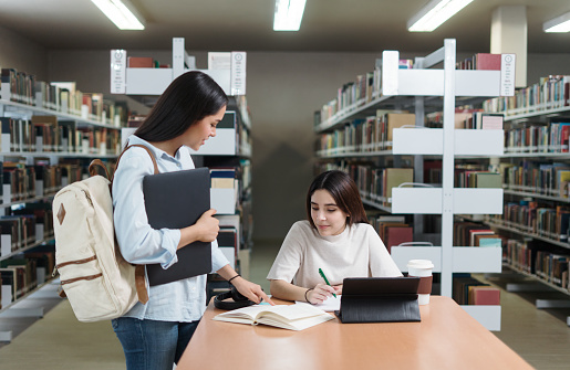 A latin female student helping a friend to study and pointing on a book with her finger.