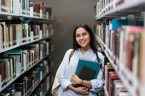A happy female student leaning on bookshelves at the library and smiling at the camera.